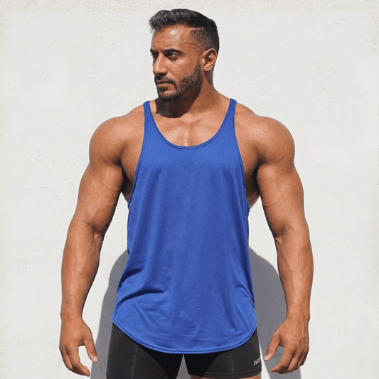 A muscular man wearing a royal blue athletic tank top and black shorts, standing against a light textured wall.