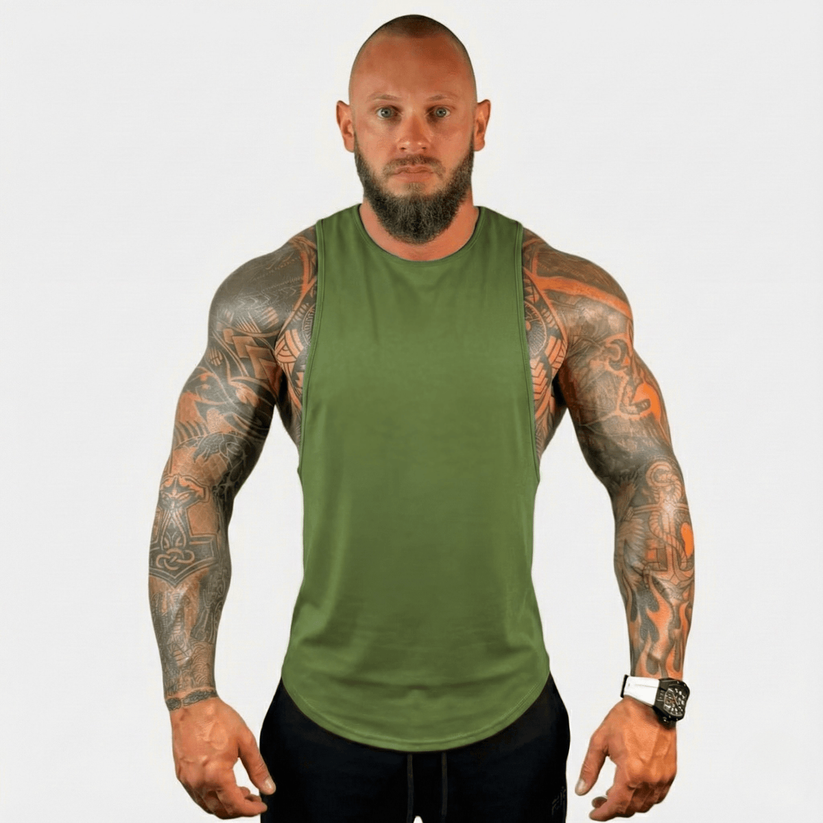 A muscular man with tattoos wearing an olive green sleeveless tank top and black pants, standing against a light gray background.