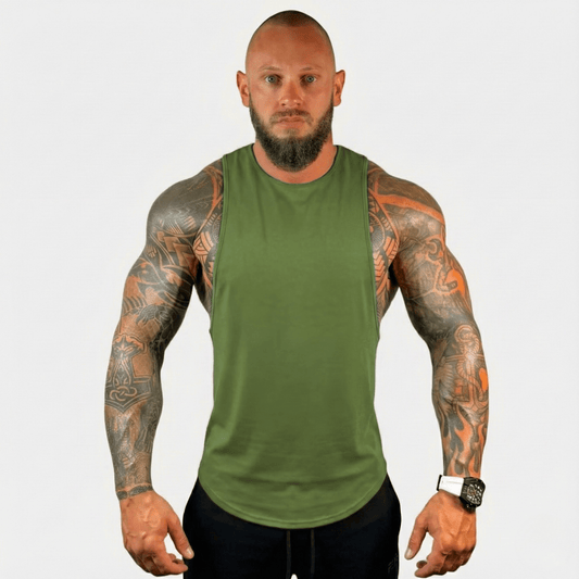 A muscular man with tattoos wearing an olive green sleeveless tank top and black pants, standing against a light gray background.