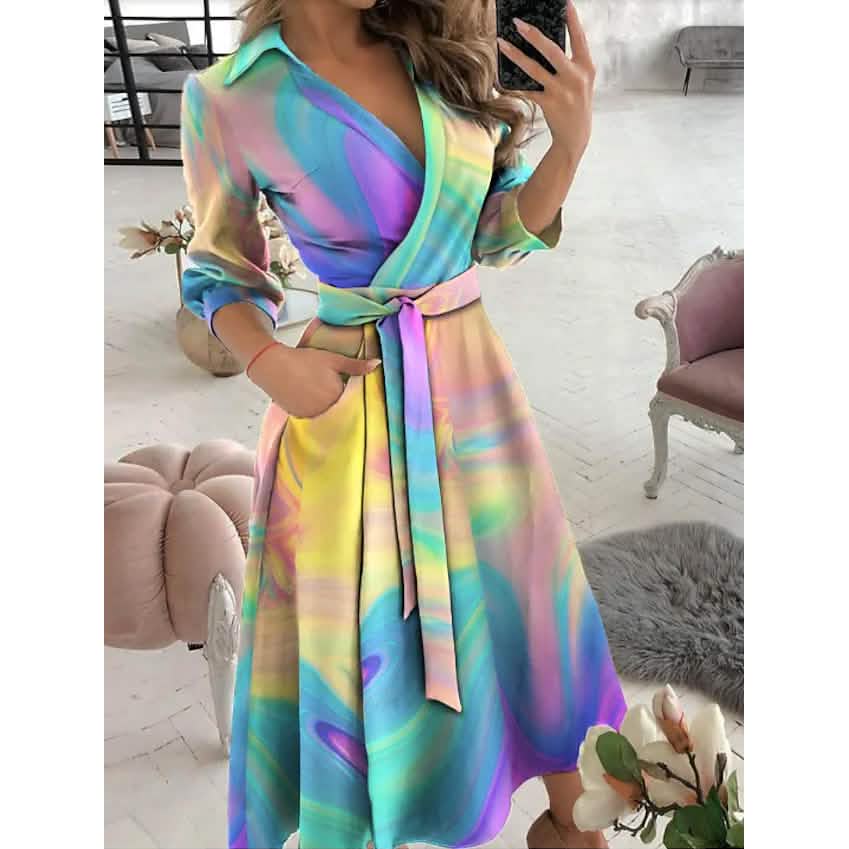 A woman takes a mirror selfie wearing a long-sleeve wrap-style midi dress with a tie waist and an abstract pastel rainbow print. The dress features blended shades of pink, yellow, blue, green, and purple.
